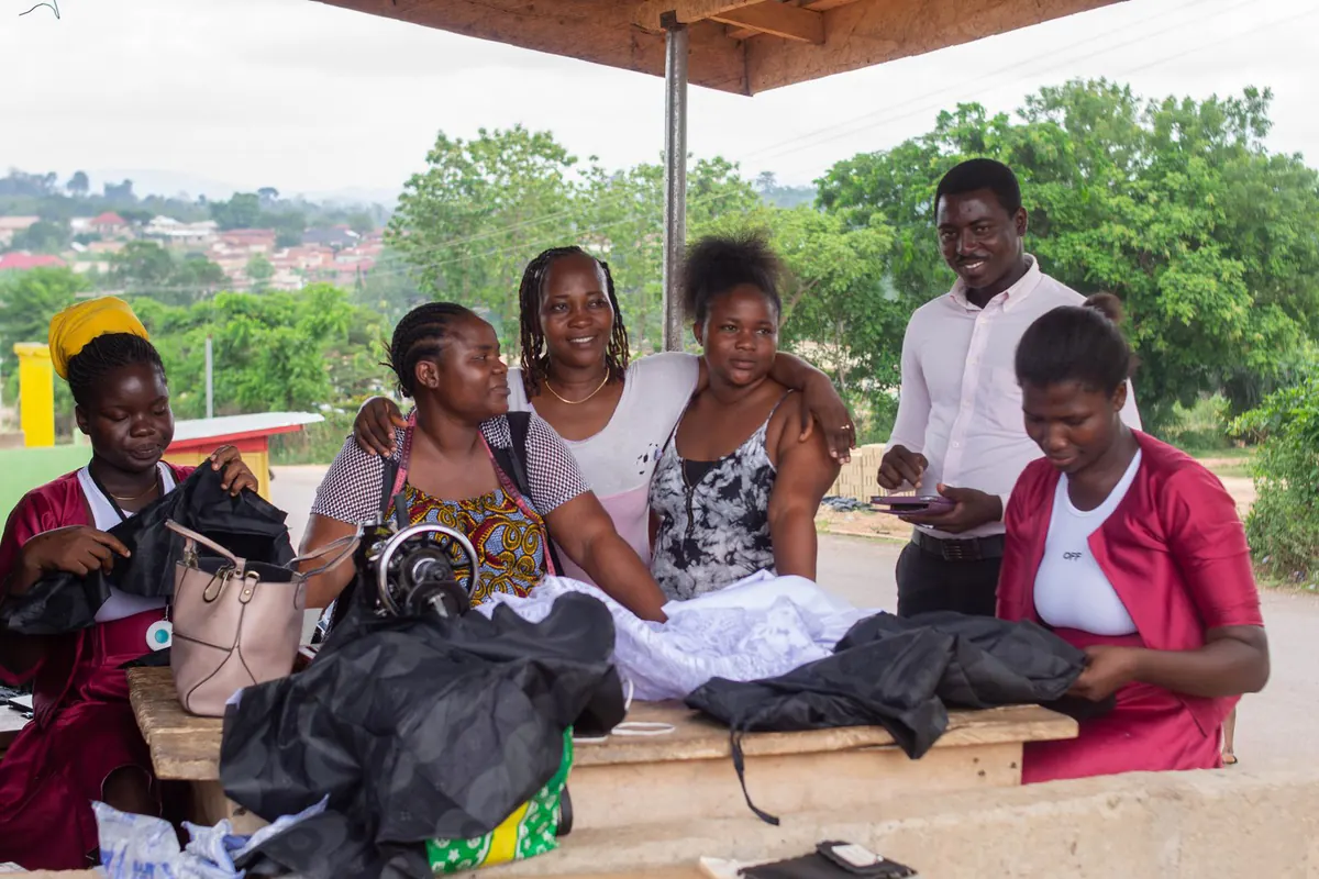 Young women in a tailoring training group with their finished garments, alongside a field researcher