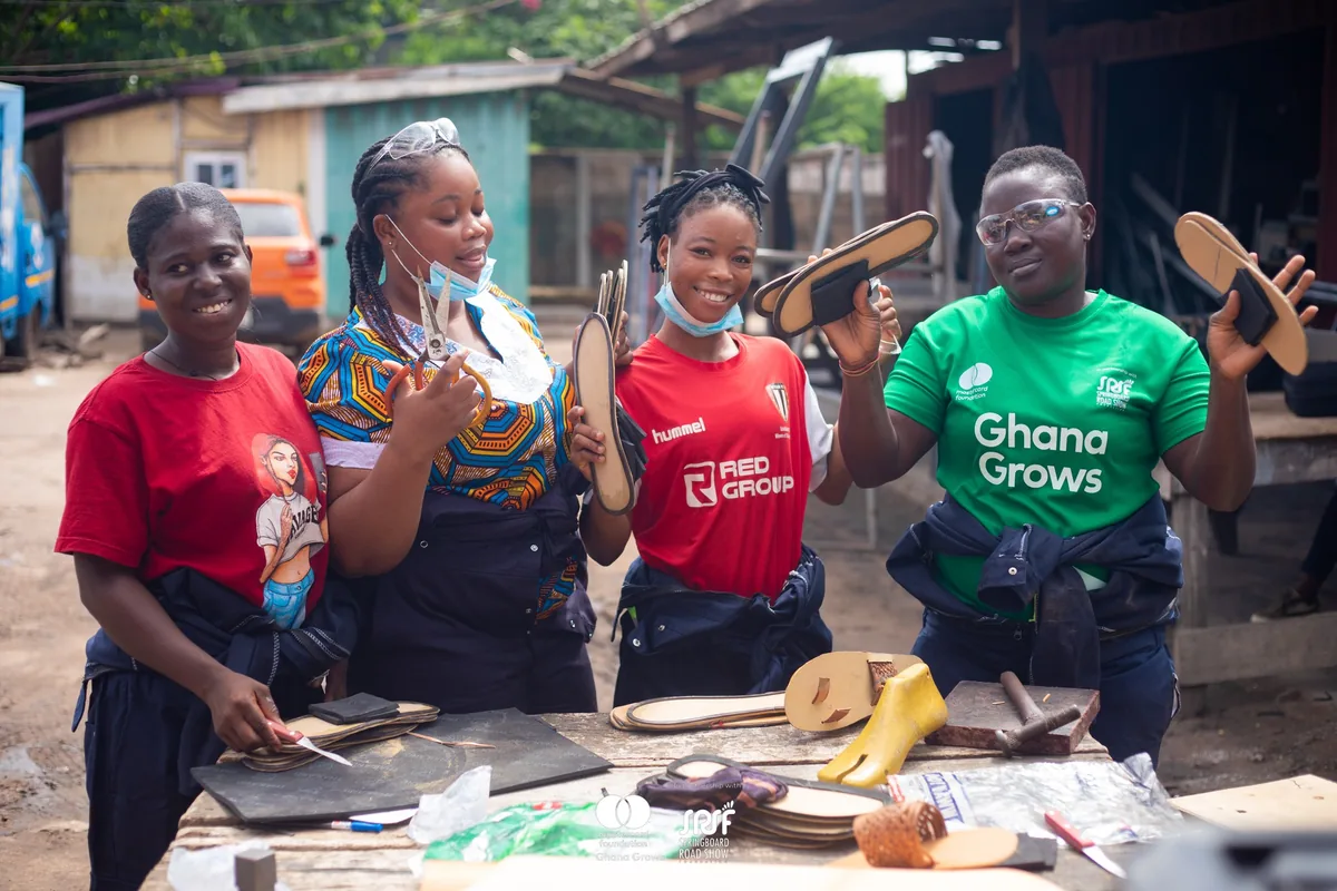 Four young women in a footwear workshop holding up shoe soles they have cut, one wearing a Ghana Grows shirt